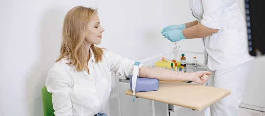 Młoda kobieta siedzi w gabinecie medycznym i wyciąga rękę do pobrania krwi. Obok stoi pracownik medyczny w jednorazowych rękawiczkach, przygotowujący się do zabiegu. Na stoliku znajdują się prob&oacute;wki laboratoryjne.