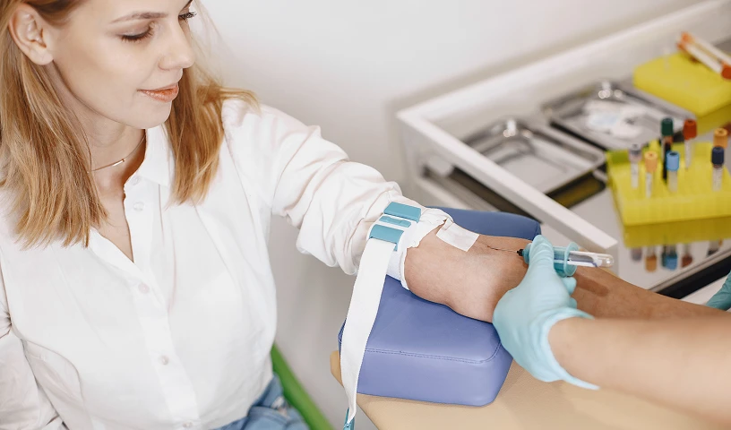 Zbliżenie na rękę pacjentki podczas pobierania krwi &mdash; na przedramieniu założona jest opaska uciskowa, a dłonie pracownika medycznego w rękawiczkach wykonują zabieg.